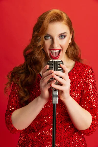 Portrait of beautiful Caucasian woman, soloist singing in microphone isolated over red background