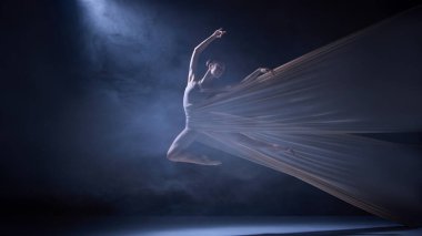 Bej kumaşa sarılı balerin sahne ışığı altında yüksek atlama yapıyor. Sahne sanatları görselleri için fiziksel disiplin, koreografi hassasiyeti ve yaratıcı hareket kavramı.