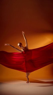 Kırmızı kumaşlı kadın balerin sıcak ışıkta dengeli poz veriyor. Modern dans ve tasarım projeleri için sanatsal denge, hassasiyet ve fiziksel zarafet kavramı.