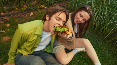 Oğlan hamburgerden büyük bir ısırık alırken, kız onu yerken gülümsüyor. Sokak yiyecekleri reklamcılığı, sosyal yaşam tarzı, gençlerin içeriği ve açık hava markaları için bağlantı kavramı.