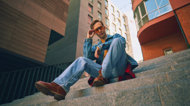 Merdivenlerde oturan genç adam kot pantolon ve turuncu gözlük takıyor. Kentsel yaşam tarzı, gençlik modası, eğitim dersleri, refah kültürü ve sosyal medya varlığı kavramı.