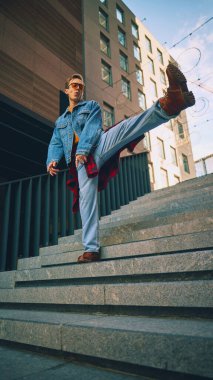 Şehir merdivenlerinde kot ceketli ve turuncu gözlüklü şık bir adam bacağını tekmeliyor. Sokak kıyafetleri, müzik alt kültürleri, yaşam tarzı, kentsel reklamcılık ve sosyal medya içeriği kavramı.