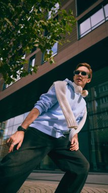 Kulaklıklı mavi kapüşonlu adam cam bina ve ağaçların yanında yürüyor. Seyahat yaşam tarzı kavramı, modern teknoloji, sokak modası, kişisel refah, yaratıcı markalaşma.