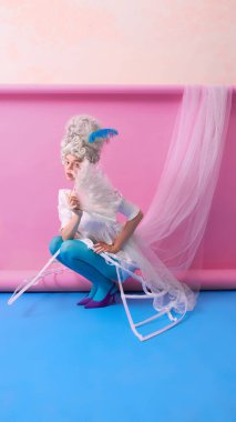 Rococo kostümlü genç bir kadın pembe arka planda tüylü bir yelpaze ile neşeyle çömeliyor. Moda tasarımı kavramı, medya tanıtımı, sosyal ağ markalaşması ve eğitici görsel.