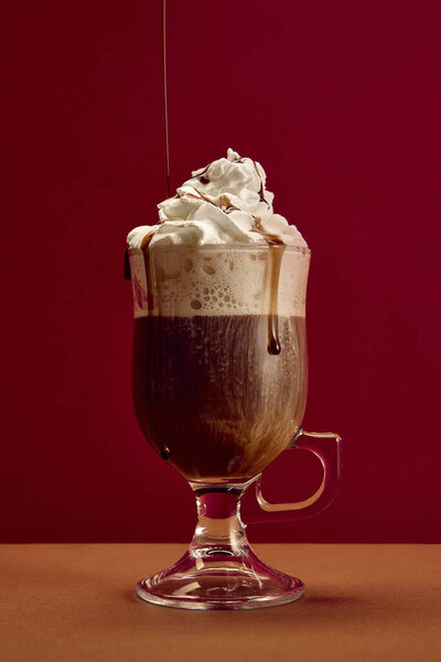 Coffee with whipped cream and chocolate syrup drizzle in clear glass against red background. Concept of dessert beverage, festive indulgence, cafe branding, and sweet luxury lifestyle.