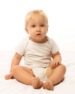 Yumuşak beyaz bir yatakta oturan sevimli bebek parlak doğal ışıkta meraklı ve sakin görünüyor. Erken çocukluk kavramı, masumiyet, bakım ve aile yaşam tarzı tasviri.