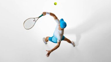Parlak mavi üniformalı tenisçi yukarıdan beyaz stüdyo katında yakalanan servisi hazırlıyor. Dijital tasarım kavramı, yaratıcı düzen, spor estetiği ve kompozisyon referansı.