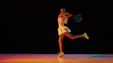 Genç tenisçi enerjik ters vuruş sırasında parlak ışıklarla aydınlatılıyor. Sporda tutku kavramı, harekette yaratıcılık ve modern atletik ifade.