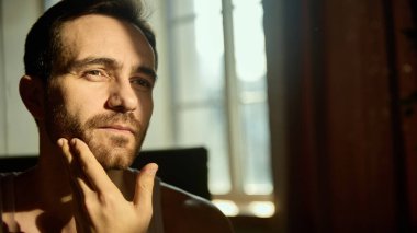 Kendine güveni tam bir adam sabah güneşinde tenini kontrol ediyor. Marka yetiştiren erkekler, berber dükkanı görselleri, cilt bakımı reklamları, sağlık platformları, günlük bakım estetiği.