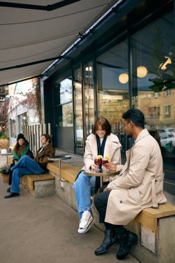 Soğuk sonbahar öğleden sonrasında modern kafenin dışında ılık içecekler paylaşan bir çift. Mevsimlik içecek tanıtımı, samimi buluşma hikayeleri, konukseverlik markaları ve atmosfer görselleri.