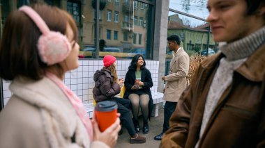 Genç arkadaşlar kış verandasındaki koltuklarda sıcak içeceklerle rahatlıyorlar. Seyahat ve şehir keşfi kavramı, günlük kahve kültürü tanıtımı, mobil dağıtım yaşam tarzı ve pozitif refah iletişimi..