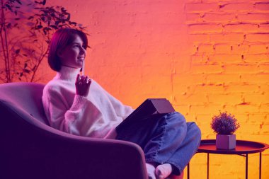 Tablet tabletli ve neon ışıklı arka planda rahatlayan bir kadın. Konfor verimliliği, uzaktan okuma, gelişigüzel öğrenme, çevrimiçi platformlar, bloglar ve pazarlama için görsel varlıklar kavramı.