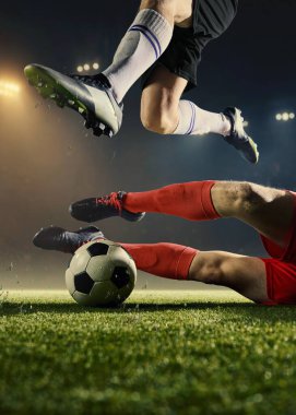 Gece stadyumundaki sahada top ve bacaklarla futbol kayma anı. Savunma oyunu, maç yoğunluğu, rekabetçi düello, spor reklamcılığı, profesyonel atletizm kavramı.