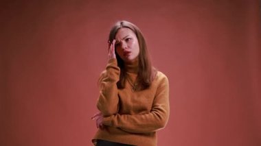 Genç bir kadın, kahverengi stüdyo arka planında düşünceli bir ifadeyle yüzüne dokunuyor. Karar verme kavramı, tereddüt, problem çözme, seçim hakkındaki endişe, yansıtıcı duygusal tepki.