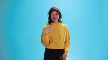 Neşeli gülüşü olan genç bir kadın mavi stüdyo arka planındaki saçlara dokunuyor. Flört kavramı, cazibe, hafif romantik ilgi, sosyal etkileşim, neşeli ruh hali, kendinden emin kadınsı ifade.