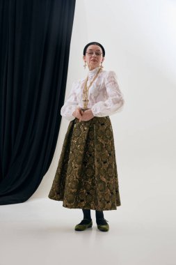 Şair özlü dantelli bluz ve desenli etek giymiş zarif olgun bir kadın. Klasik moda portresi konsepti, tam giysi sunumu, yaş kapsayıcı tarz, editoryal ve ticari tasvirler.