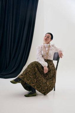 Klasik stüdyo ortamında sandalyede oturan zarif, klasik stil bir kadın. Zamansız kadınlık kavramı, retro moda estetiği, zarif duruş, kültürel miras tarzı, editoryal ve tasarım kullanımı.