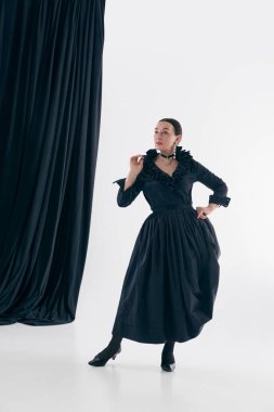 Stüdyoda siyah elbiseli bir kadın perdenin yanında poz veriyor. Moda performansı kavramı, klasik kadınlık, şair özü atmosfer, editoryal poz, zamansız ifade.