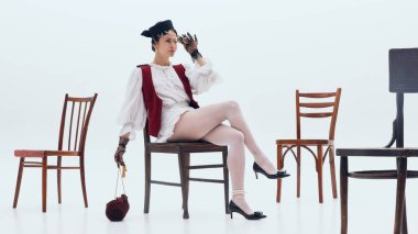 Minimalist beyaz alanda klasik dürbünle oturan şık bir kadın. Moda editörü, marka anlatımı tasviri, butik tanıtım görselleri ve sanatsal reklam kompozisyonu kavramı.