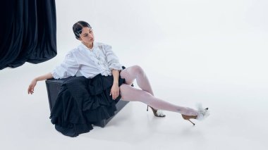 Katmanlı etek ve beyaz bluz minimalist stüdyo içinde siyah kutuya yaslanmış bir kadın. Şair modası, sanatsal içerik, lüks markalar hikaye anlatımı ve modern gardırop sunumu kavramı.