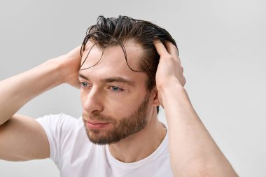 Beyaz tişörtlü yetişkin sakallı bir adamın önünde iki eliyle ıslak saçlar tutuyordu. Alnına iplikler düşüyordu. Açık gri stüdyo arka planı vardı..
