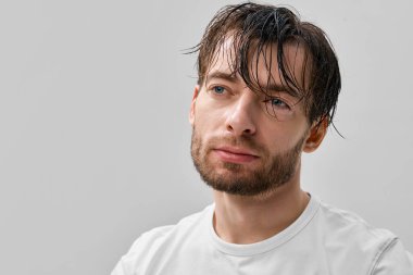 Islak saçlı, alnında beyaz t-shirt olan açık gri minimal stüdyo arkaplanı olan yetişkin kısa sakallı bir adamın ön portresi..