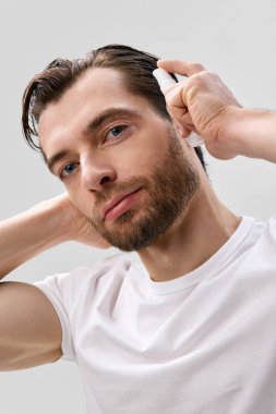 Beyaz tişörtlü, yetişkin sakallı bir adam kıvırcık saçın yanında küçük bir sprey sprey şişesi tutuyor, başını hafifçe çevirmiş, kameraya bakıyor, açık gri stüdyo arka planında.