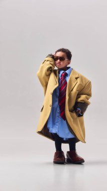 Küçük çocuk büyük beden ceket kravat takıyor ve güneş gözlüğü takıyor. Stüdyoda güven içinde poz veriyor. Liderlik hayalleri, çocukluk hırsı, moda rolü oynama ve gelecekteki meslekler hakkında hayal gücü kavramı.