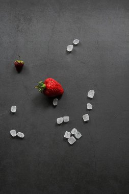Koyu arkaplanda çilek, iki küçük çilek, kırmızı meyve, etrafa saçılmış büyük şeker kristalleri, beyaz şeker
