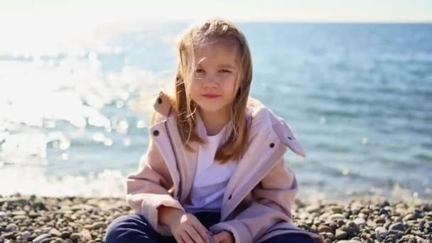 Een klein meisje in een jas zit op een kiezelstrand — Stockvideo