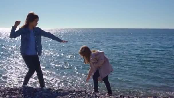 Twee meisjes lopen op een kiezelstrand en gooien stenen in het water — Stockvideo