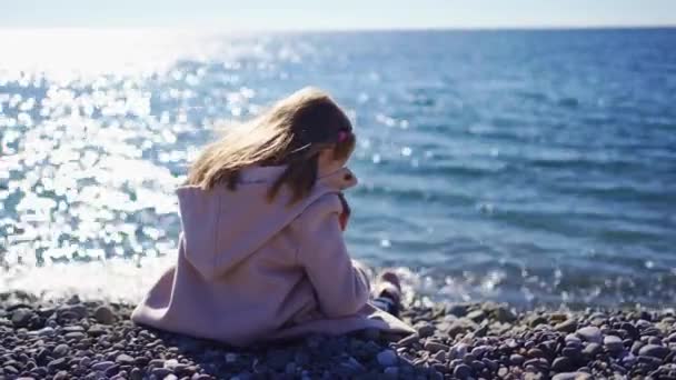Little girl in a coat on a pebble seashore. walking by the sea to restore health — Stock Video