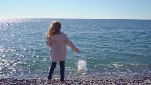 Lány séta a kavicsos strandon, és dobja köveket a vízbe — Stock videók