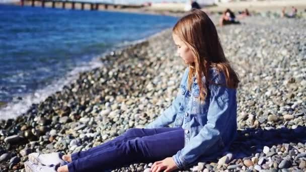 Een meisje gooit stenen in het water en grappen terwijl ze op een strand zit. — Stockvideo