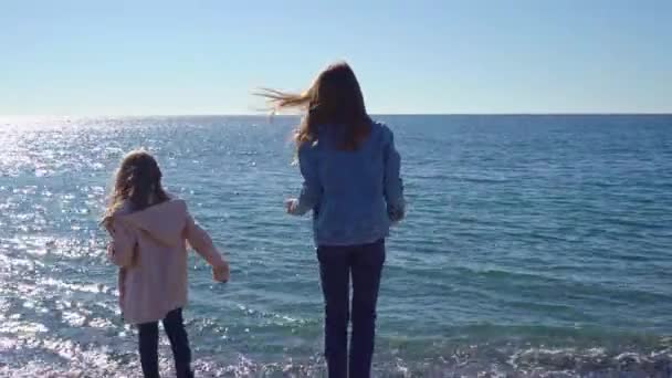 Δύο κορίτσια περπατούν σε μια παραλία με βότσαλα και πετάνε πέτρες στο νερό. — Αρχείο Βίντεο