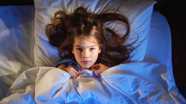 Vista de cima. Uma menina está na cama, assustada e cobre o rosto com um cobertor. — Vídeo de Stock