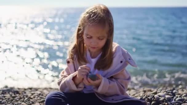 Een meisje in een jas zit op een kiezelstrand en schildert met een steen op een steen. — Stockvideo