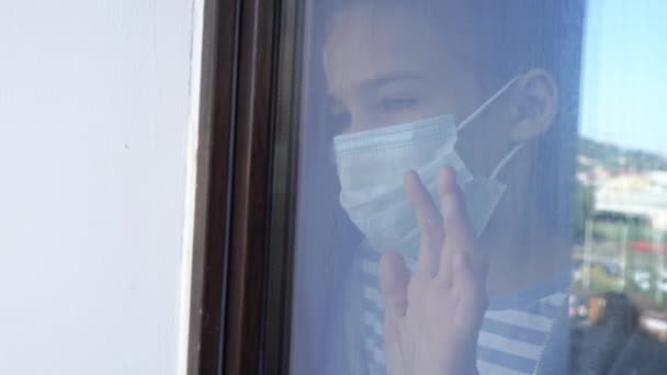Genom glas. tonåring flicka i skyddsmask, titta ut genom fönstret utanför. — Stockvideo