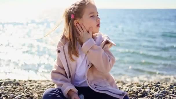 Uma menina em um casaco senta-se em uma costa do mar de seixos e tosse — Vídeo de Stock