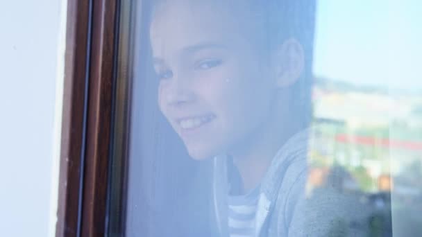 Přes sklo. šťastná dospívající dívka se dívá z okna ven. obnoveno. — Stock video