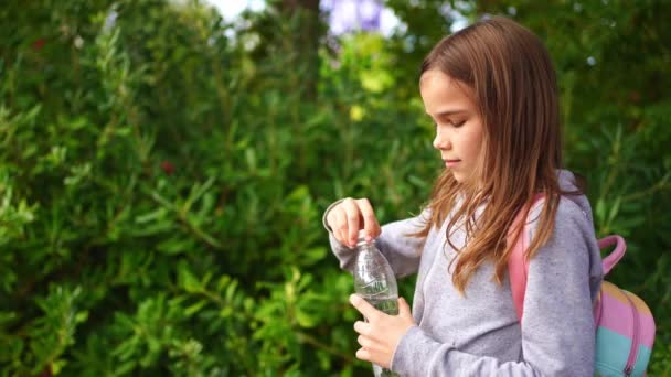 Una chica desenrosca la tapa de una botella de agua bebiendo mientras camina por el parque. — Vídeo de stock