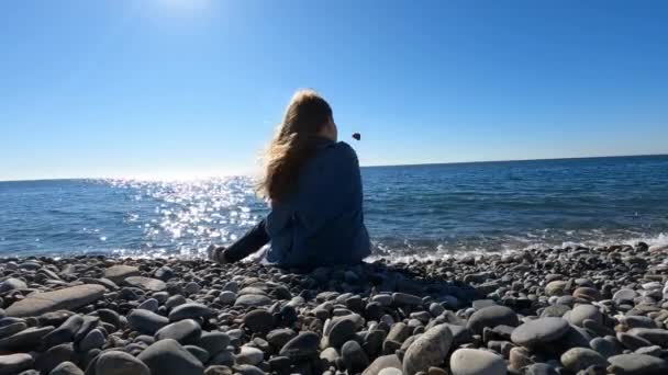 Zadní pohled. dívka hodí kameny do vody a vtipy, zatímco sedí na břehu moře. — Stock video