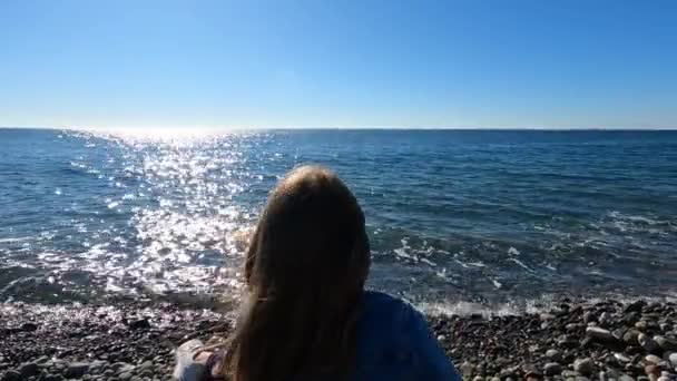 Visão traseira. menina joga pedras na água e piadas enquanto sentado na costa do mar. — Vídeo de Stock