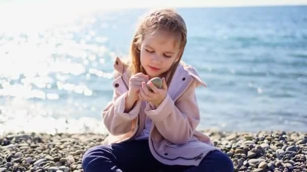 En flicka i rock sitter på en stenstrand och målar med en sten på en sten. — Stockvideo