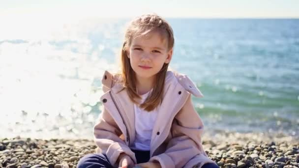 Ένα κοριτσάκι με παλτό κάθεται σε μια βοτσαλωτή ακτή. — Αρχείο Βίντεο