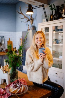 a young woman drinks hot cocoa sitting on the kitchen table. Christmas pastries.
