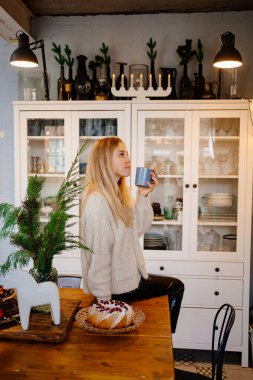 a young woman drinks hot cocoa sitting on the kitchen table. Christmas pastries.