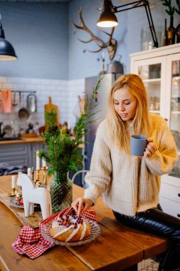 a young woman drinks hot cocoa sitting on the kitchen table. Christmas pastries.