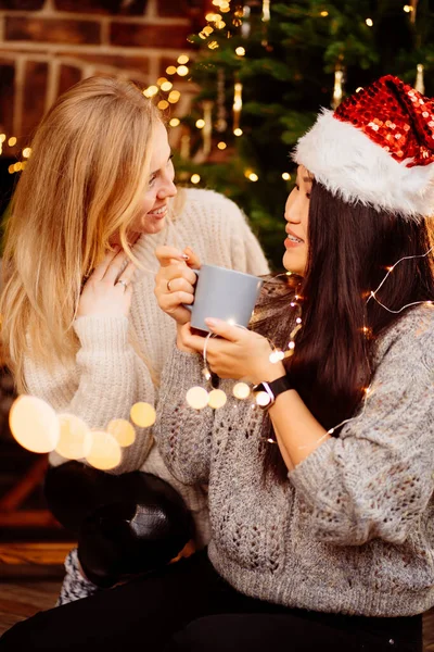 Two woman drinking tea by fireplace with New Years decoration. Romantic.