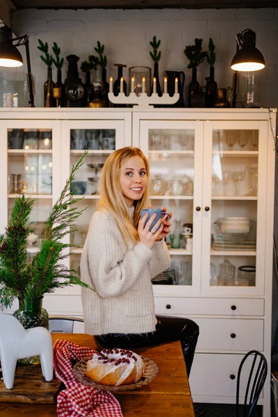 a young woman drinks hot cocoa sitting on the kitchen table. Christmas pastries.